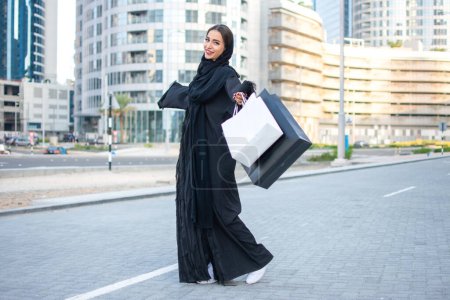 Téléchargez les photos : Heureuse femme arabe en tenue traditionnelle tenant des sacs à provisions dans la rue de la ville - en image libre de droit