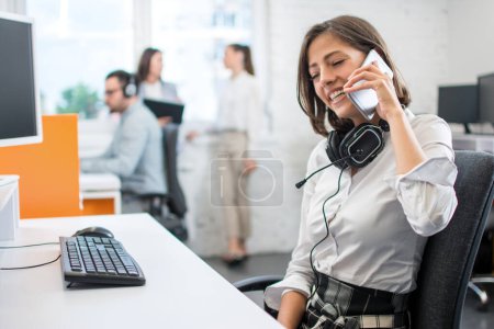 Téléchargez les photos : Jeune femme souriante en tenue de travail et casque parlant sur téléphone portable alors qu'elle était assise sur son lieu de travail au bureau. - en image libre de droit
