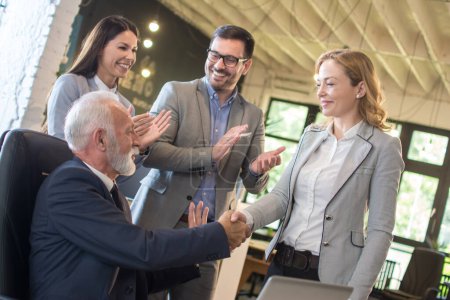 Senior boss and middle aged female employee shaking hands with colleagues clapping hands around them in the office