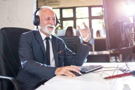Téléchargez les photos : Homme d'affaires senior avec casque utilisant l'ordinateur dans le bureau - en image libre de droit