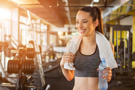 Foto de Hermosa mujer sonriente en forma joven con toalla y botella de agua preparándose para el entrenamiento deportivo en el gimnasio. - Imagen libre de derechos