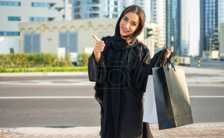 Foto de Joven mujer árabe hermosa vistiendo abaya y sosteniendo una bolsa de compras mientras camina por la calle. Mujer árabe feliz haciendo como signo con los dedos. - Imagen libre de derechos