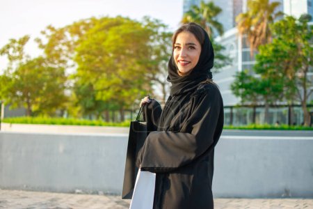 Téléchargez les photos : Jeune femme musulmane qui aime faire du shopping dans la ville d'Abu Dhabi. Belle femme arabe avec des sacs à provisions dans la rue. - en image libre de droit