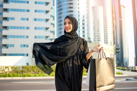 Photo for Young beautiful Arabic woman wearing abaya and holding a shopping bags while walking on street - Royalty Free Image
