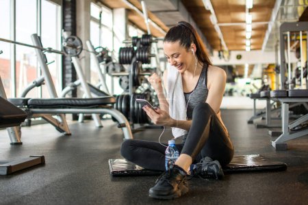 Téléchargez les photos : Joyeux jeune femme lisant de bonnes nouvelles sur le téléphone mobile et célébrant le succès pendant la pause d'entraînement dans la salle de gym. - en image libre de droit