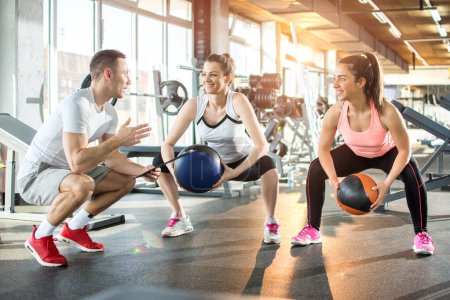 Téléchargez les photos : Deux femmes sportives faisant des exercices avec des balles de fitness avec l'aide de leur entraîneur personnel dans la salle de gym - en image libre de droit