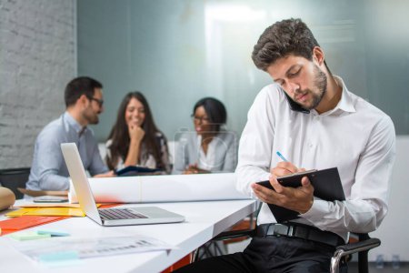 Young businessman holding mobile phone between ear and shoulder talking and writing notes to clipboard in modern office, while his business partners having meeting in the background.
