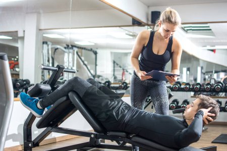 Téléchargez les photos : Beau homme faisant assis sur le banc de poids avec l'aide d'instructeur personnel féminin attrayant à la salle de gym - en image libre de droit