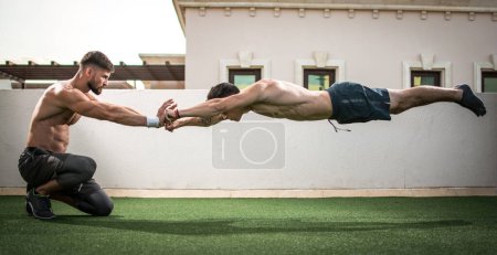 Foto de Hombre musculoso guapo haciendo flexiones con saltos con la ayuda de entrenador personal en el gimnasio de la azotea - Imagen libre de derechos