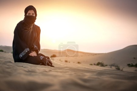 Foto de Mujer joven beduina vestida con ropa negra tradicional contra la puesta del sol en el cielo sobre el desierto - Imagen libre de derechos