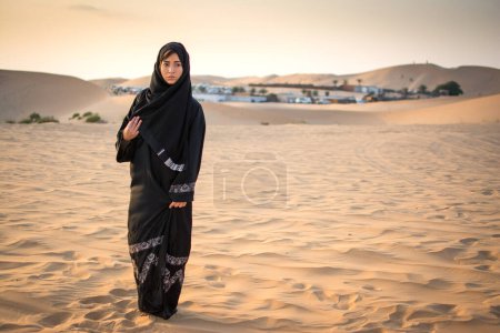 Foto de Retrato de larga duración de una mujer árabe vestida con ropa negra tradicional parada en el desierto frente al pueblo beduino - Imagen libre de derechos