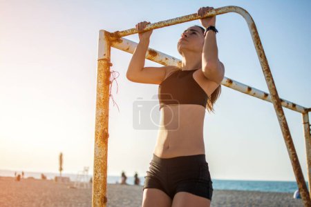 Photo for Sporty fit young woman doing pull ups on metal goal frame on sandy beach during sunset - Royalty Free Image