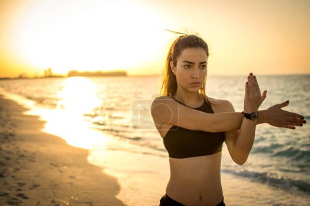 Photo for Portrait of concentrated fit young sportswoman warming up at the beach during sunset - Royalty Free Image