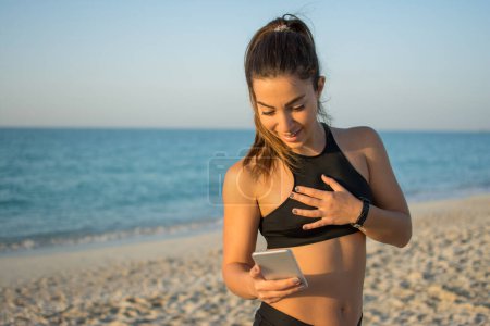 Foto de Mujer deportiva en forma sorprendida usando el teléfono durante su entrenamiento en la playa - Imagen libre de derechos