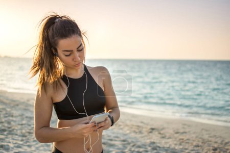 Photo for Portrait of young woman with earphones listening to music and text messaging on smart phone at the beach - Royalty Free Image