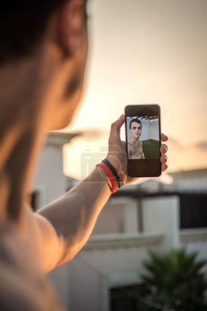 Foto de Vista por encima del hombro de los hombres jóvenes sosteniendo el teléfono inteligente y tomando un selfie al aire libre durante la puesta del sol - Imagen libre de derechos