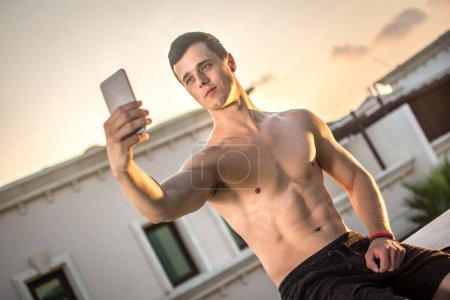 Foto de Hombre joven musculoso guapo sin camisa tomando selfie con teléfono inteligente mientras está sentado en la azotea durante el atardecer - Imagen libre de derechos