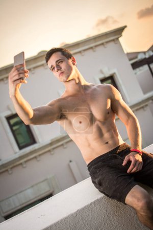 Foto de Hombre musculoso guapo sin camisa tomando una selfie mientras está sentado en la pared de los tejados al aire libre durante el atardecer - Imagen libre de derechos