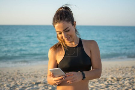 Téléchargez les photos : Souriant fitness jeune femme utilisant le téléphone à la plage - en image libre de droit