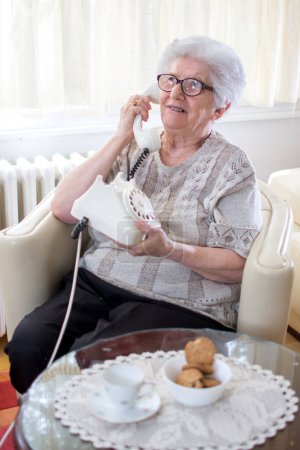 Foto de Sonriente anciana hablando por teléfono fijo giratorio mientras se relaja en el sillón en casa - Imagen libre de derechos