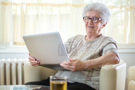 Téléchargez les photos : Femme âgée souriante utilisant un ordinateur portable à la maison - en image libre de droit