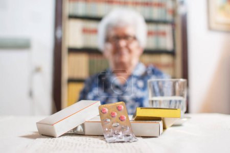 Téléchargez les photos : Gros plan de médicaments avec verre d'eau sur la table avec une femme âgée floue assise sur la chaise en arrière-plan - en image libre de droit