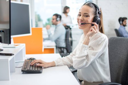 Foto de Amistoso operador de centro de llamadas dama en auriculares inalámbricos hablando con el cliente en la oficina - Imagen libre de derechos