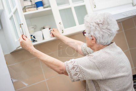 Téléchargez les photos : Vue arrière de la femme âgée prenant la vaisselle du placard dans la cuisine - en image libre de droit