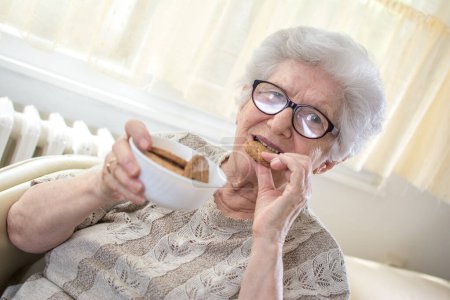 Foto de Mujer mayor con plato de galletas comiendo deliciosa galleta en casa - Imagen libre de derechos