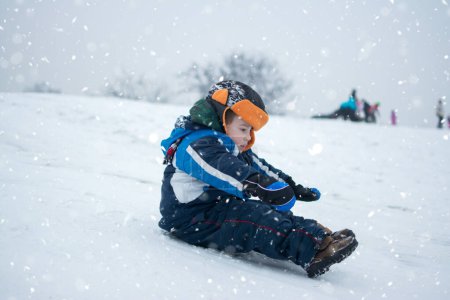 Photo for Cute little boy sledding down a hill on cold snowy on winter day - Royalty Free Image