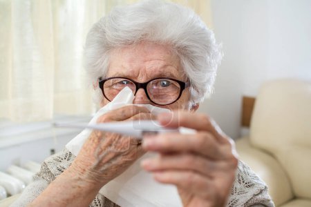 Photo for Close up portrait of sick senior woman checking body temperature with thermometer and holding a tissue paper over her face. - Royalty Free Image