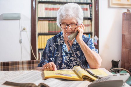 Photo for Senior woman talking on phone and searching yellow pages for services - Royalty Free Image