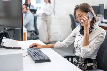 Zufriedene Kundenbetreuerin mit Headset am Handy an ihrem Arbeitsplatz im Callcenter-Büro