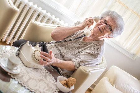 Foto de Sonriente abuela hablando por teléfono fijo giratorio mientras está sentado en el sillón en casa - Imagen libre de derechos