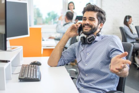 Glücklicher männlicher Kundendienstmitarbeiter mit Headset, der in der Büropause auf dem Handy spricht
