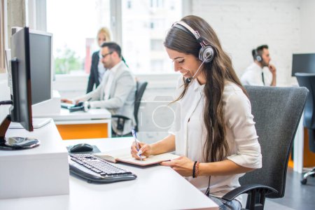 Photo for Smiling woman operator agent with headset taking notes while talking with client in call center - Royalty Free Image
