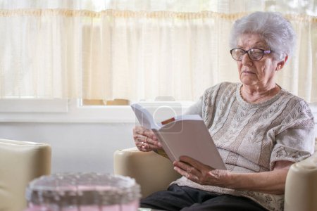 Foto de Anciana leyendo un libro mientras está sentada en la sala de estar en casa - Imagen libre de derechos