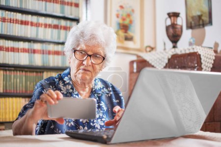 Concepto de tecnología, edad y personas. Mujer mayor usando smartphone y laptop en casa