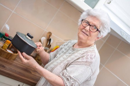 Téléchargez les photos : Femme âgée va réchauffer le repas dans une casserole prise au réfrigérateur. Grand-mère tenant la casserole dans la cuisine. Femme âgée souriante préparant la nourriture. - en image libre de droit