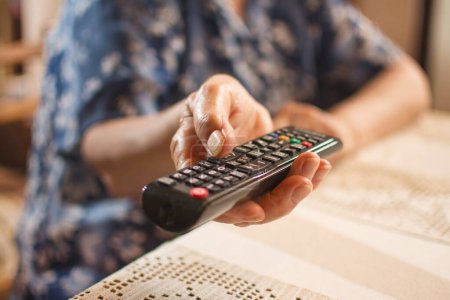 Photo for Close up of senior woman sitting on chair near the desk and holding tv remote control. Selective focus - Royalty Free Image