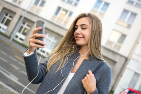 Foto de Sonriente chica rubia con smartphone escuchando música en la calle de la ciudad - Imagen libre de derechos