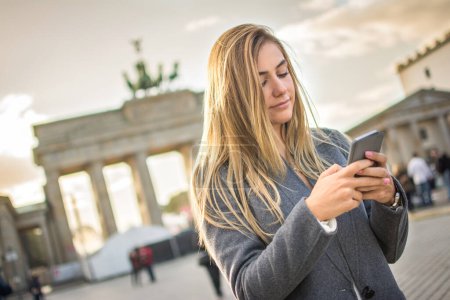 Téléchargez les photos : Belle fille aux cheveux blonds utilisant le téléphone devant la porte de Brandebourg à Berlin, Allemagne - en image libre de droit