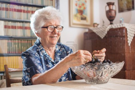 Photo for Smiling old woman eating grapes at home - Royalty Free Image