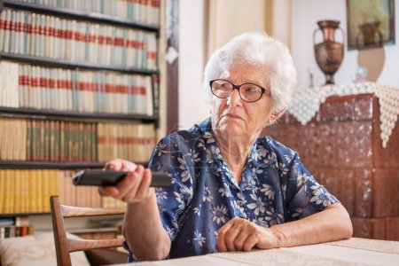 Téléchargez les photos : Heureuse femme âgée regarder la télévision et changer de chaîne avec télécommande à la maison - en image libre de droit