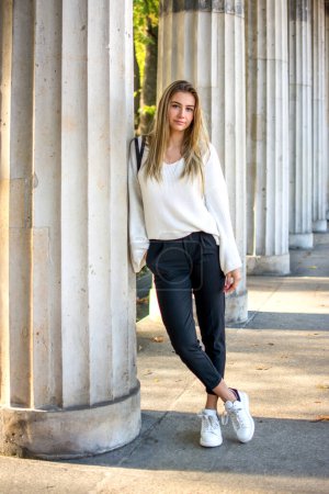 Full lenght of young woman in fashion clothing leaning against pillar outdoors