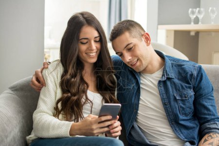 Foto de Feliz pareja joven mirando juntos el teléfono inteligente mientras están sentados en el sofá en casa - Imagen libre de derechos