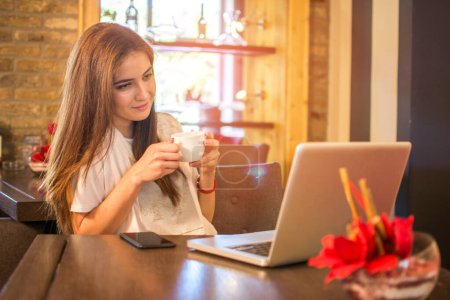 Téléchargez les photos : Adolescente prendre une tasse de café et apprendre sur ordinateur portable dans un café - en image libre de droit