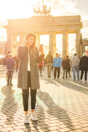 Téléchargez les photos : Toute la longueur de la jeune femme à l'écoute de la musique sur téléphone portable tout en se tenant près de la porte Brandebourg à Berlin - en image libre de droit