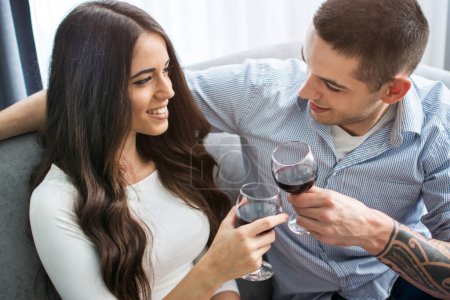 Foto de Hermosa joven pareja amorosa sentada cerca la una de la otra y beber vino tinto en casa - Imagen libre de derechos
