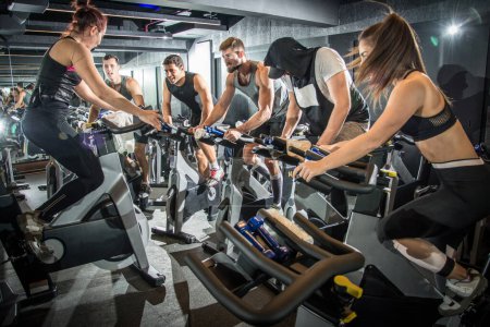 Téléchargez les photos : Groupe de personnes de remise en forme qui font de l'entraînement cardio sur les vélos d'intérieur à la salle de gym - en image libre de droit